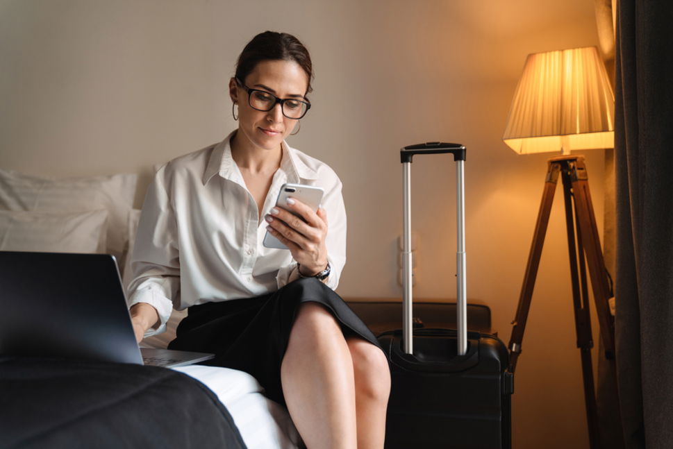 Woman with suitcase sitting on hotel bed using laptop and smartphone